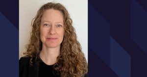 Portrait de Karine Barrette avec les cheveux longs frisés châtains, yeux bleu et un rire pincé. Chandail noir.