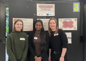 Trois étudiantes en Techniques de la documentation qui posent fièrement devant leur kiosque lors de de la présentation de leur projet.