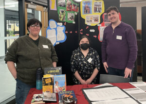 Deux étudiantes et un étudiant en Techniques de la documentation qui posent fièrement devant leur kiosque lors de de la présentation de leur projet.