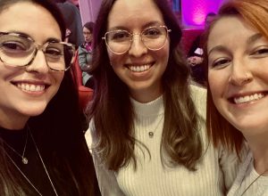Trois étudiante en avant-plan souris à la caméra dans la salle où se déroule le Gala.