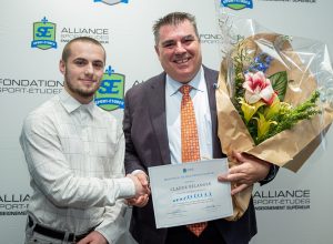 François-Xavier Béland et Claude Bélanger pose devant lelogo de l'Alliance sport-études avec certificat et gros bouquet de fleurs dans les mains.