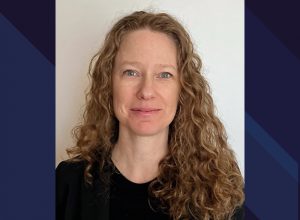 Portrait de Karine Barrette avec les cheveux longs frisés châtains, yeux bleu et un rire pincé. Chandail noir.