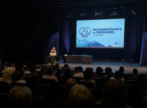 Foule assise dans l'ausitorium dans la pénombre vue de dos avec la directrice éclairée au lutrin avec un grand écran sur la scène sur laquelle on peut lire Reconnaissance du personnel